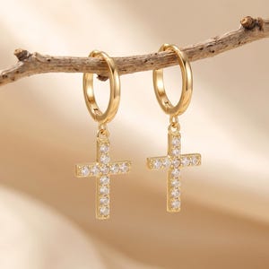 May include: A pair of gold hoop earrings featuring dangling cross charms. Each cross is encrusted with small, clear, sparkling stones. The earrings are suspended from a thin, natural wooden twig, showcasing a polished metallic finish.