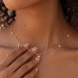 May include: A delicate gold chain necklace featuring five evenly spaced, small, round, clear stone charms. The necklace rests against the collarbone, showcasing a thin, polished metal link design with a secure clasp mechanism visible at the back.