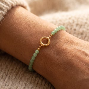 May include: A beaded bracelet featuring small, round, light green stones strung together. The bracelet is secured with a circular gold-toned metal clasp positioned at the centre, connecting the two ends of the beaded strand with small gold beads.