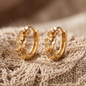 Könnte beinhalten: Ein Paar goldene Creolen, die mit einer Reihe kleiner, miteinander verbundener Blumenformen auf der Vorderseite verziert sind. Jede Blume ist in der Mitte mit einem winzigen, klaren, funkelnden Stein besetzt, was diesen runden Ohrringen ein filigranes Aussehen verleiht.