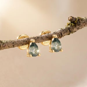 Puede incluir: Un par de pendientes de botón dorados con piedras de ágata musgosa en forma de lágrima con inclusiones verdes visibles. Cada pendiente tiene un engaste dorado con el sello S925, que indica una base de plata de ley, sobre una rama de madera.