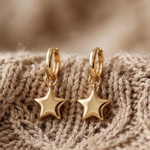 May include: A pair of gold-toned hoop earrings, each featuring a polished, five-pointed star charm suspended from a small circular hoop. The earrings have a smooth, reflective metallic finish and a simple, clean design with no additional embellishments.