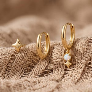 May include: A set of gold-toned jewelry consisting of a four-pointed star stud earring, a plain gold hoop earring, and a gold hoop earring featuring a dangling white opal-like stone set above a small four-pointed star charm.