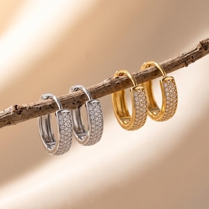 May include: Two pairs of small hoop earrings are displayed on a thin wooden branch. One pair is silver-toned and the other is gold-toned. Each hoop features a flat front surface densely encrusted with tiny, clear, sparkling stones in a pavé setting.