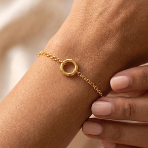 Op de afbeelding: Een goudkleurige kettingarmband met een centrale cirkelvormige ringbedel. De armband is opgebouwd uit fijne, uniforme ronde schakels die verbonden zijn met de gladde, gepolijste gouden cirkel in het midden voor een eenvoudig ontwerp.
