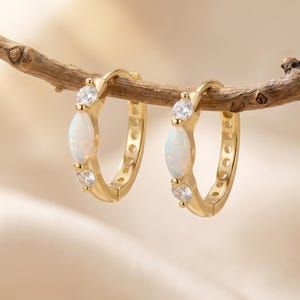 May include: A pair of gold hoop earrings featuring a central marquise-cut opal stone flanked by two small round clear stones on each hoop. The earrings are suspended from a thin, textured brown twig, showcasing the polished gold metal finish.