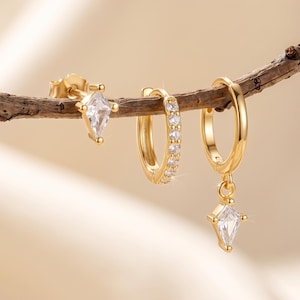 May include: A set of three gold-toned earrings featuring a kite-shaped stud with a clear stone, a small hoop earring encrusted with tiny clear stones along the front, and a gold hoop earring with a dangling kite-shaped charm containing a clear stone.