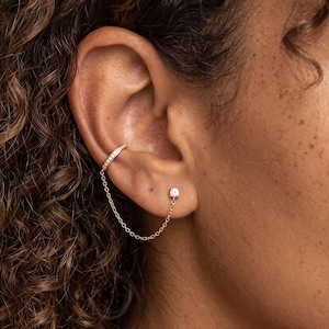 May include: A gold-toned ear cuff featuring a row of small clear stones, connected by a delicate fine chain to a single stud earring with a clear stone. The jewelry is worn on the cartilage and the earlobe, creating a linked chain design.
