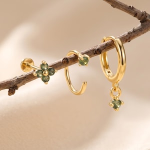 May include: Three gold-tone earrings featuring green stones. One is a flat-back stud with a four-petal flower design, one is a small open hoop with a single stone, and one is a hinged hoop with a dangling stone charm. Each piece is marked with S925.