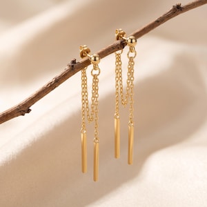 May include: A pair of gold-toned earrings featuring a stud post with a small sphere, from which two delicate chains hang, each ending in a slender, polished gold-toned bar. The earrings are suspended from a thin, brown wooden twig.