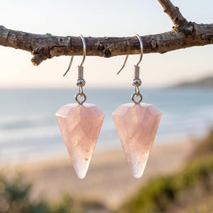 May include: A pair of dangle earrings featuring faceted, cone-shaped rose quartz gemstones suspended from silver-toned metal fishhook wires. The translucent, pale pink stones have a geometric, multi-faceted cut that tapers to a point.