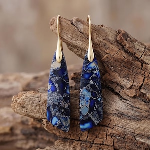 Puede incluir: Un par de pendientes colgantes con piezas triangulares alargadas que presentan un patrón de mosaico en tonos azul profundo, azul marino y fragmentos de plata metálica, suspendidos de ganchos de metal en tono dorado.