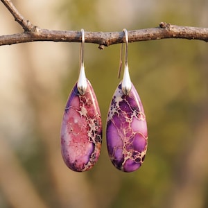Puede incluir: Un par de pendientes en forma de lágrima con una piedra veteada en tonos morados y rosas. Los pendientes cuelgan de ganchos plateados. El patrón de la piedra es único. El fondo es borroso y natural.