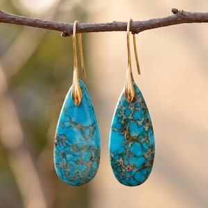 May include: A pair of turquoise teardrop earrings with gold-colored hooks. The earrings are hanging from a brown branch. The turquoise stones have a unique pattern of brown and gold veining.