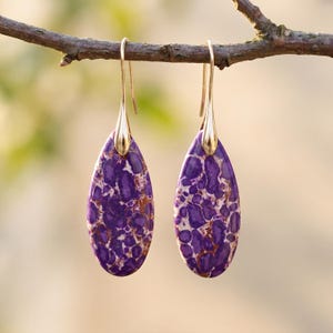 May include: A pair of teardrop-shaped earrings with a gold-colored hook and a purple and white speckled stone. The earrings are hanging from a tree branch against a blurred background.