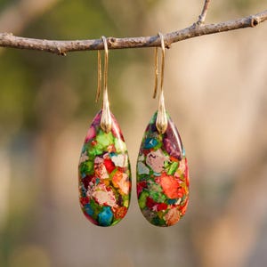 May include: A pair of teardrop-shaped earrings with gold-colored hooks. The earrings feature a mosaic of colors including red, green, blue, and purple. The earrings are hanging from a small branch.