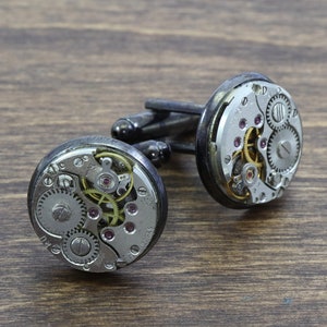 May include: A pair of silver cufflinks with exposed clockwork mechanisms. The cufflinks are on a brown wooden surface.