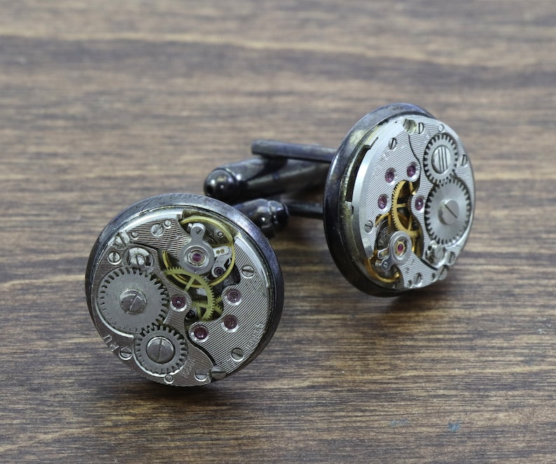 May include: A pair of silver cufflinks with exposed clockwork mechanisms. The cufflinks are on a brown wooden surface.