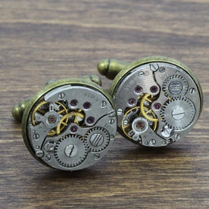 May include: A pair of gold-toned cufflinks with exposed clockwork mechanisms. The cufflinks are made of metal and have a circular shape. The clockwork is visible through the clear face of the cufflinks.