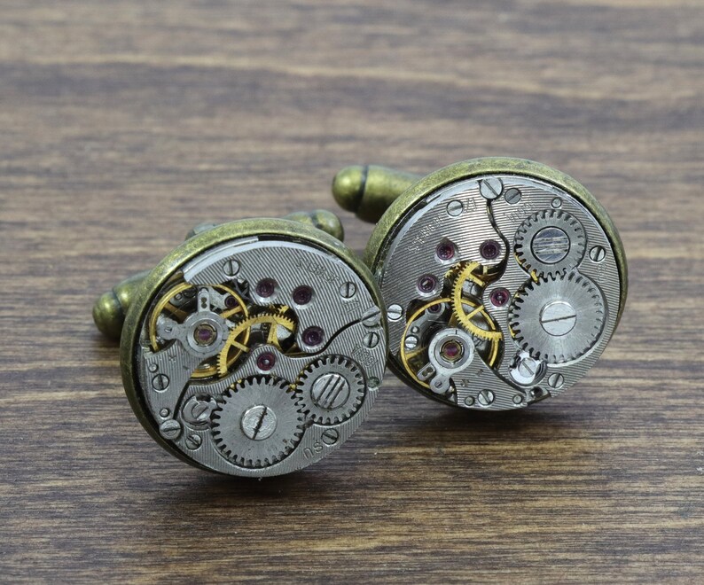 May include: A pair of gold-toned cufflinks with exposed clockwork mechanisms. The cufflinks are made of metal and have a circular shape. The clockwork is visible through the clear face of the cufflinks.