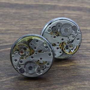 May include: Two silver-toned watch movements with exposed gears and intricate details. The watch movements are circular and have a metallic finish.