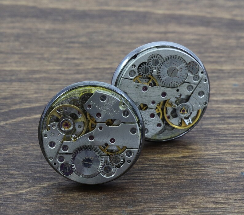 May include: Two silver-toned watch movements with exposed gears and intricate details. The watch movements are circular and have a metallic finish.