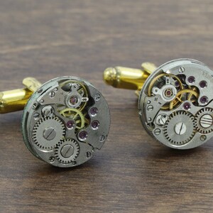 May include: A pair of silver cufflinks with gold accents. Each cufflink features a circular design with exposed watch gears and a small, clear crystal in the center.