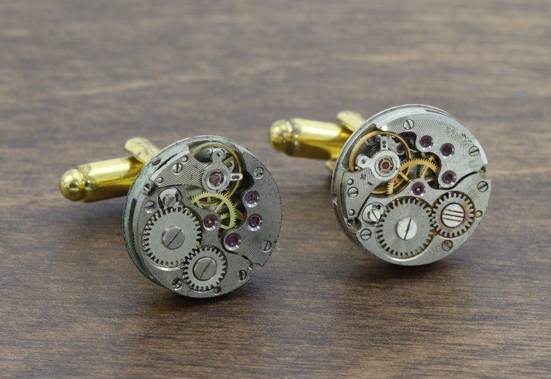 May include: A pair of silver cufflinks with gold accents. Each cufflink features a circular design with exposed watch gears and a small, clear crystal in the center.