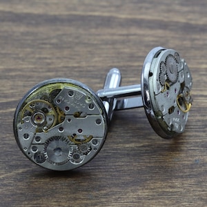 May include: Silver cufflinks with exposed clockwork mechanisms. The cufflinks are made of metal and have a silver finish. The clockwork is visible through the clear face of the cufflinks.