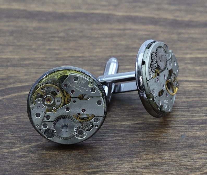 May include: Silver cufflinks with exposed clockwork mechanisms. The cufflinks are made of metal and have a silver finish. The clockwork is visible through the clear face of the cufflinks.
