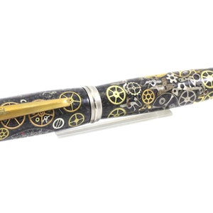 May include: A detailed pen with a steampunk design. The pen features a black barrel adorned with gold and silver gears, a gold clip with the name "SCHMMEL", and silver accents on the ends. The pen is displayed on a clear stand.