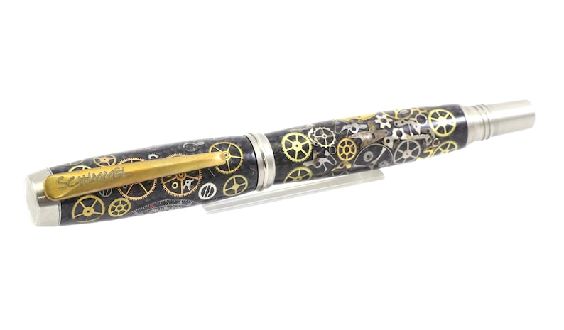 May include: A detailed pen with a steampunk design. The pen features a black barrel adorned with gold and silver gears, a gold clip with the name "SCHMMEL", and silver accents on the ends. The pen is displayed on a clear stand.