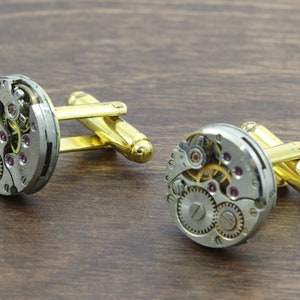 May include: A pair of gold cufflinks with a silver watch movement design. The cufflinks feature intricate gears and a small ruby-colored jewel.