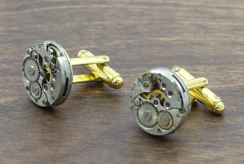 May include: A pair of gold cufflinks with a silver watch movement design. The cufflinks feature intricate gears and a small ruby-colored jewel.