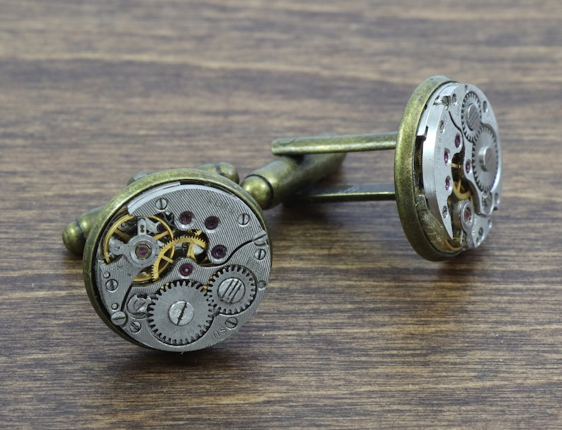May include: A pair of cufflinks with a vintage watch mechanism design. The cufflinks are made of metal with a gold finish and feature exposed gears and a silver face.