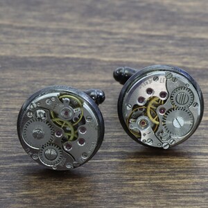 May include: Two silver cufflinks with exposed clockwork mechanisms. The cufflinks are round and have a black metal casing.