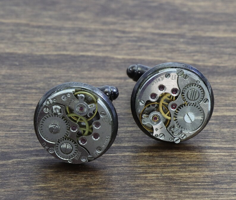 May include: Two silver cufflinks with exposed clockwork mechanisms. The cufflinks are round and have a black metal casing.