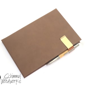 May include: A brown leather notebook with a gold metal pen holder and a pen. The notebook is closed and has a smooth surface. The pen is a brown and silver color.