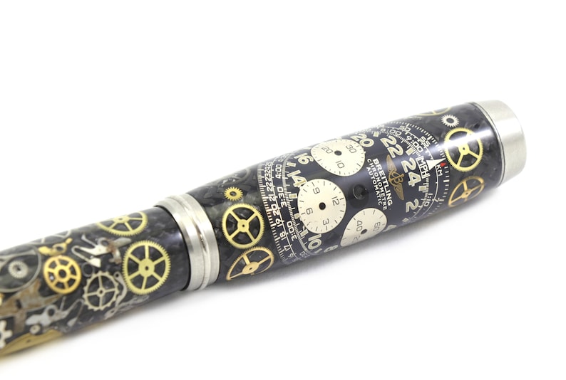 May include: A pen with a black and silver body, adorned with watch gears and clock faces. The pen features a Breitling Chronometre Automatic design with white clock faces and gold gears.