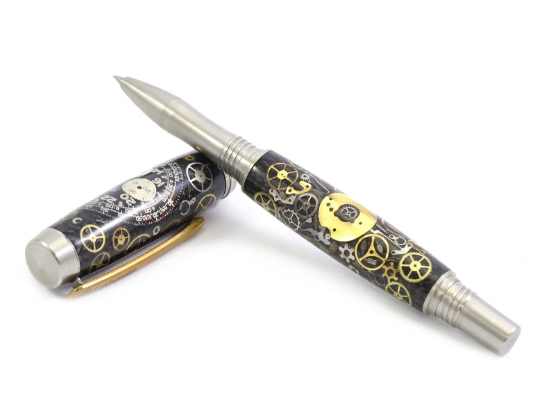 May include: A fountain pen with a stainless steel nib and accents. The pen's barrel is decorated with a pattern of gears in gold and silver tones, and the cap features a clock face design. The pen is lying on a white surface.