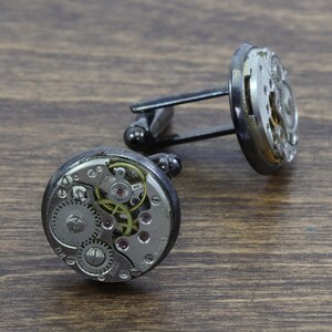 May include: A pair of silver cufflinks with exposed clockwork mechanisms. The cufflinks are attached to a black metal bar.