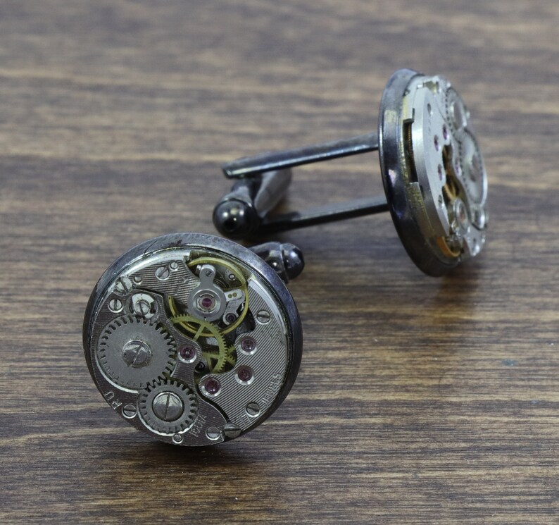 May include: A pair of silver cufflinks with exposed clockwork mechanisms. The cufflinks are attached to a black metal bar.