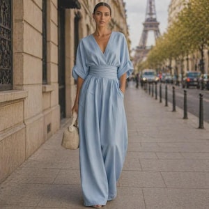 May include: A light blue jumpsuit featuring a V-neckline, wide sleeves, and a cinched waist with a wide band. The garment has long, loose-fitting trousers and is paired with a small, textured beige handbag held in one hand.