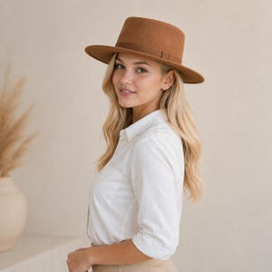 Könnte beinhalten: Ein brauner Fedora-Hut mit flachem Oberteil und einem Band um den Kronenbereich. Der Hut hat eine breite Krempe und ist aus einem filzartigen Material gefertigt. Der Hut wird von einer Person getragen, die ein weißes Hemd mit Knöpfen trägt.