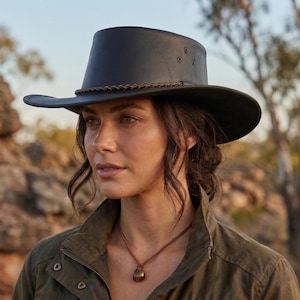 May include: A dark gray leather hat with a wide brim and a brown braided band. The hat has three small eyelets on the crown. The hat is a classic style, suitable for outdoor activities.