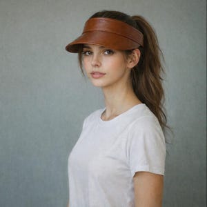 Könnte beinhalten: Braunes Leder-Visier mit gebogener Krempe. Das Visier wird auf dem Kopf getragen und soll die Augen vor der Sonne schützen. Ein stilvolles Accessoire für Outdoor-Aktivitäten.