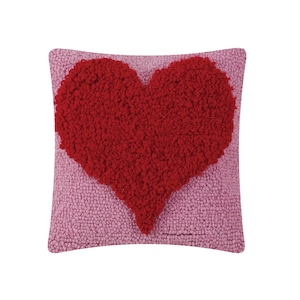 May include: A square decorative pillow with a red heart design on a pink background. The heart is textured and raised, creating a 3D effect. The pillow is likely made of a soft, plush material.