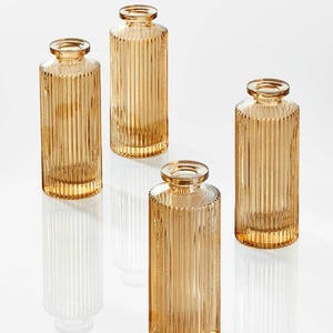 May include: Four amber-colored glass vases with vertical ribbed detailing. The vases are cylindrical with a slightly flared top and a narrow opening. The glass has a warm, golden hue, and the vases are arranged on a white surface.