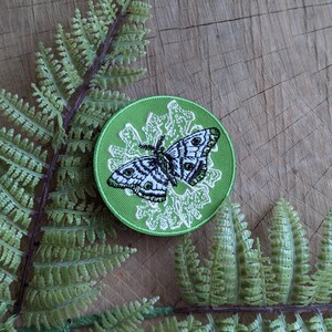 May include: A round, green embroidered patch featuring a detailed butterfly design in black and white. The butterfly is centered on a white floral pattern, all set against a bright green background. The patch is surrounded by a green border.