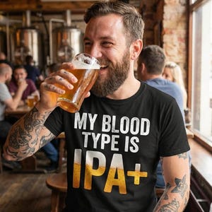Può includere: Una maglietta nera con la scritta "MY BLOOD TYPE IS IPA+" in bianco e giallo. L'immagine mostra una persona che beve birra in un bar. La persona ha tatuaggi sulle braccia e sta sorridendo.